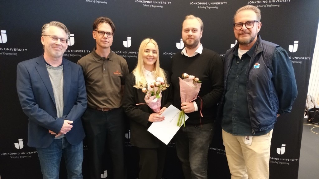 fyra män och en kvinna på bilden. Mannen i mitten och kvinnan håller i blommor och ett diplom. 