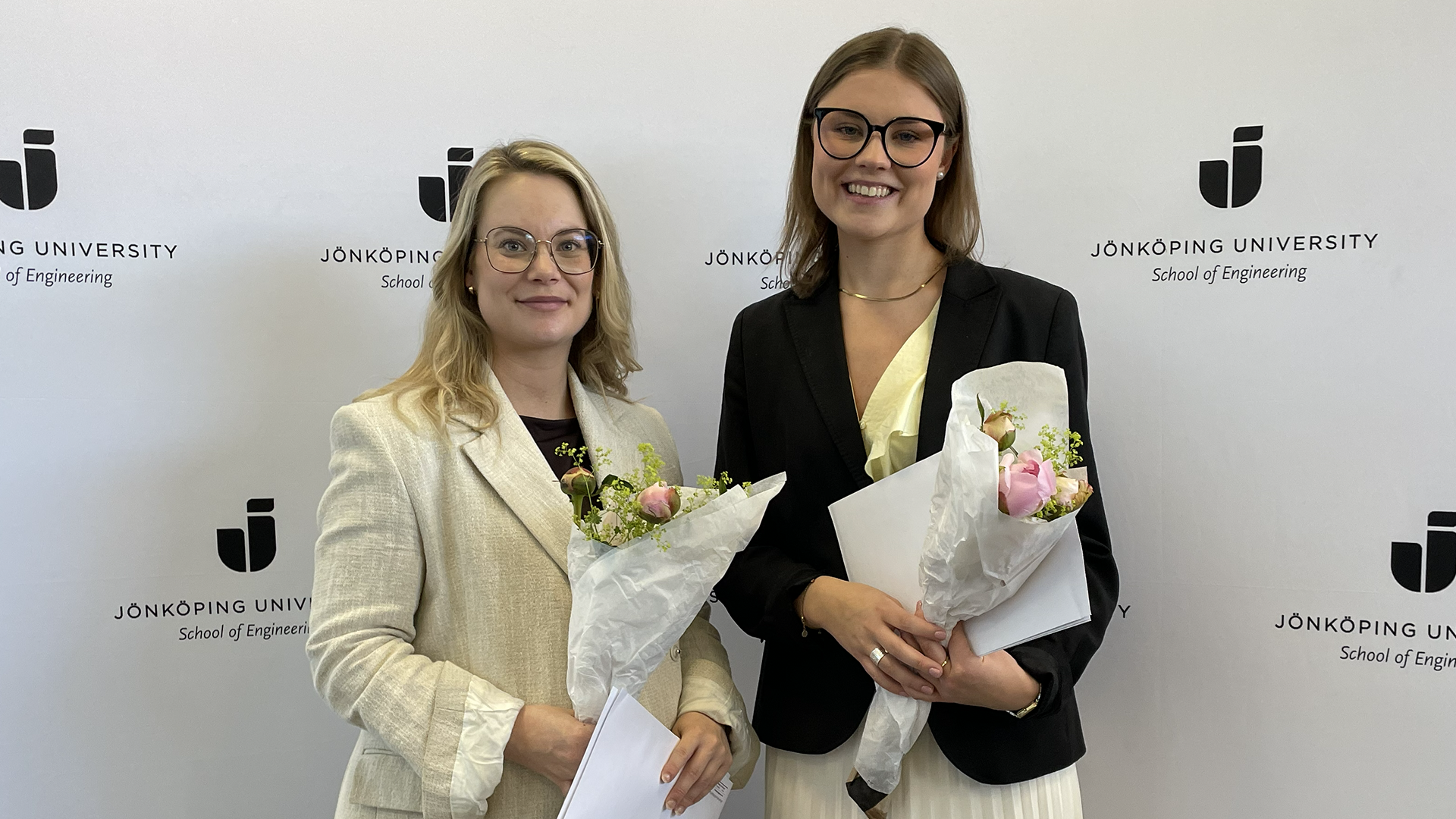 Två leende kvinnor håller varsitt diplom och blombukett.