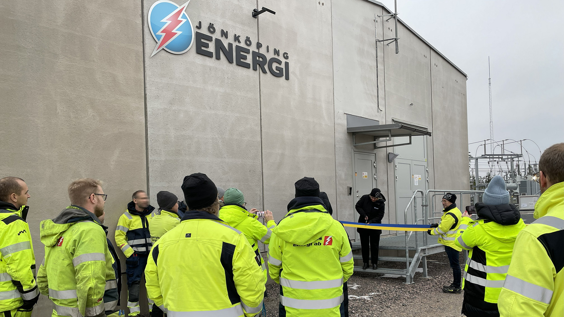 Människor med gula varseljackor tittar på när en kvinna i svarta kläder klipper ett blågult band utanför en betongbyggnad.