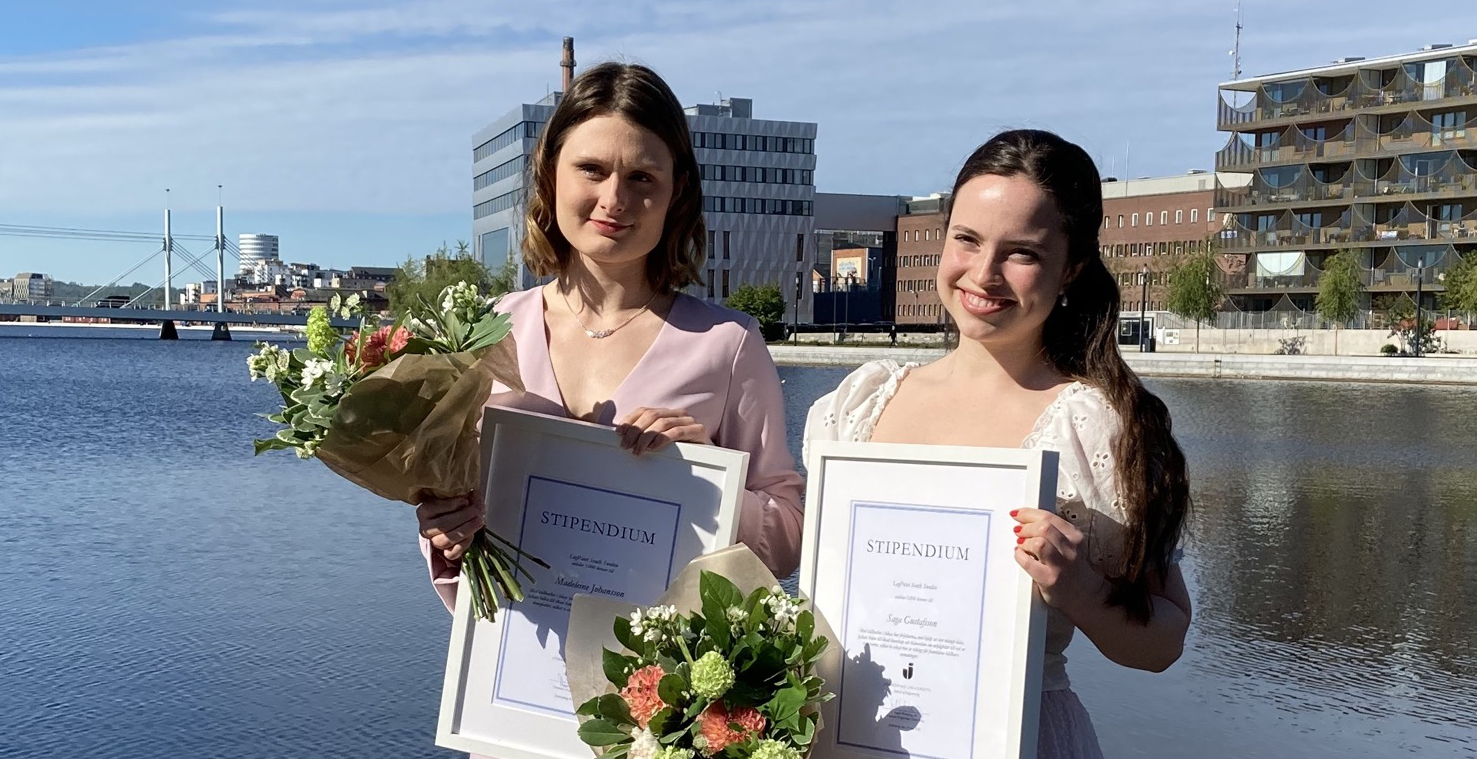 Bild på Saga och Madeleine. De är finklädda och håller i blommor och stipendium. Fotot är taget utomhus med Munksjön och en blå himmel i bakgrunden. 